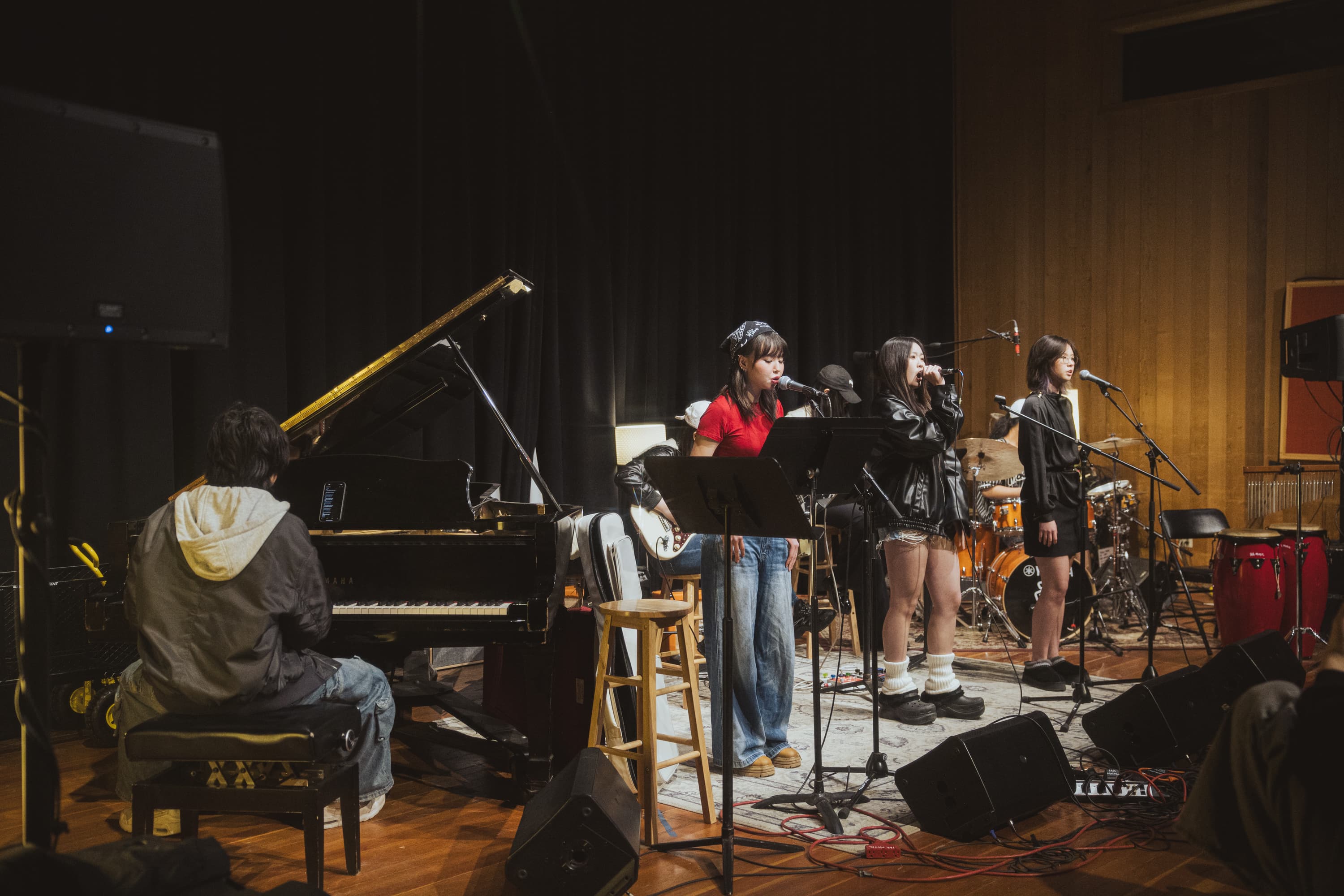 Three vocalists sing into microphones accompanied by a pianist and drummer in a music studio.
