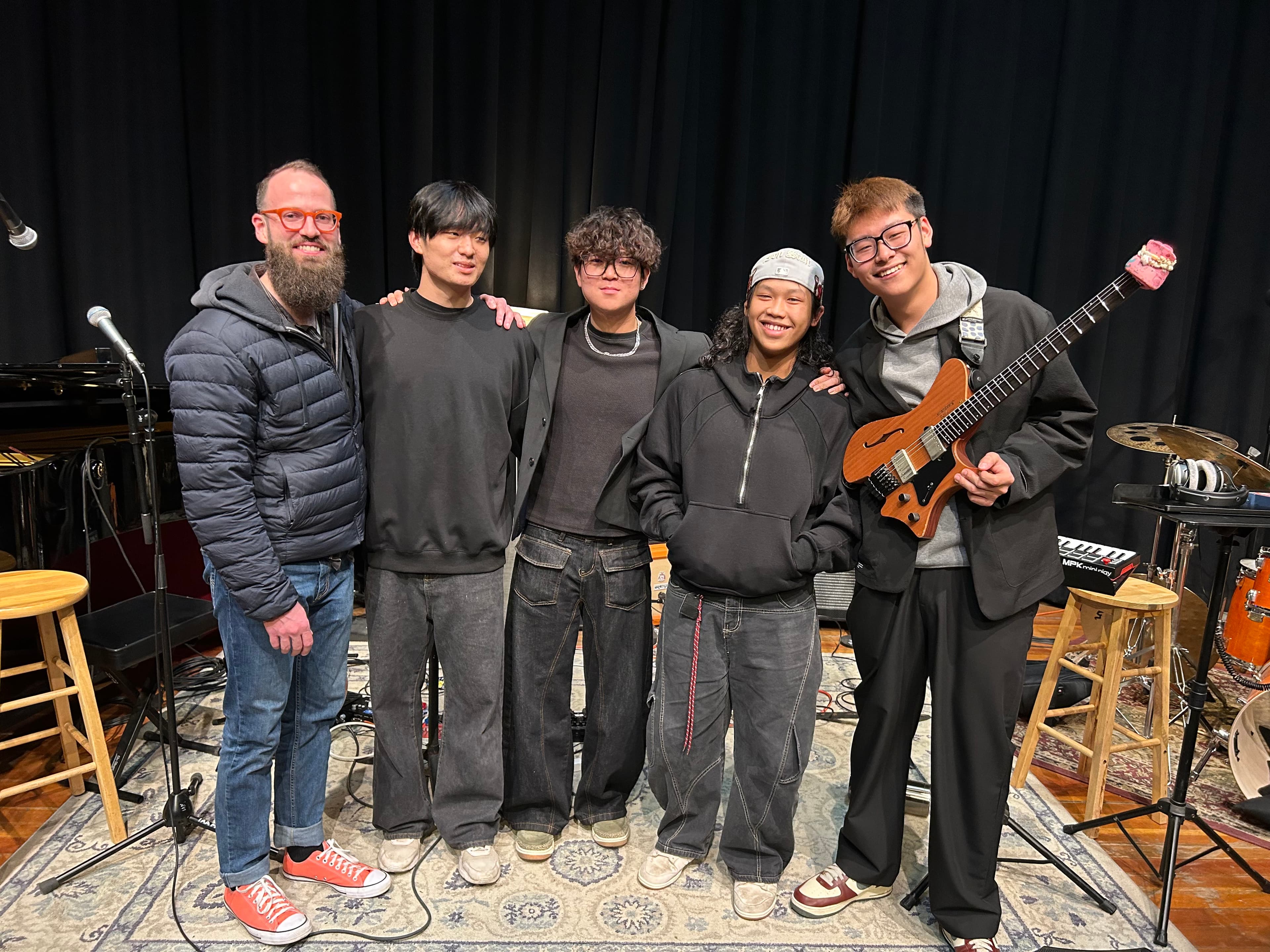 Five smiling musicians pose in a studio with a headless guitar, drums, and piano.