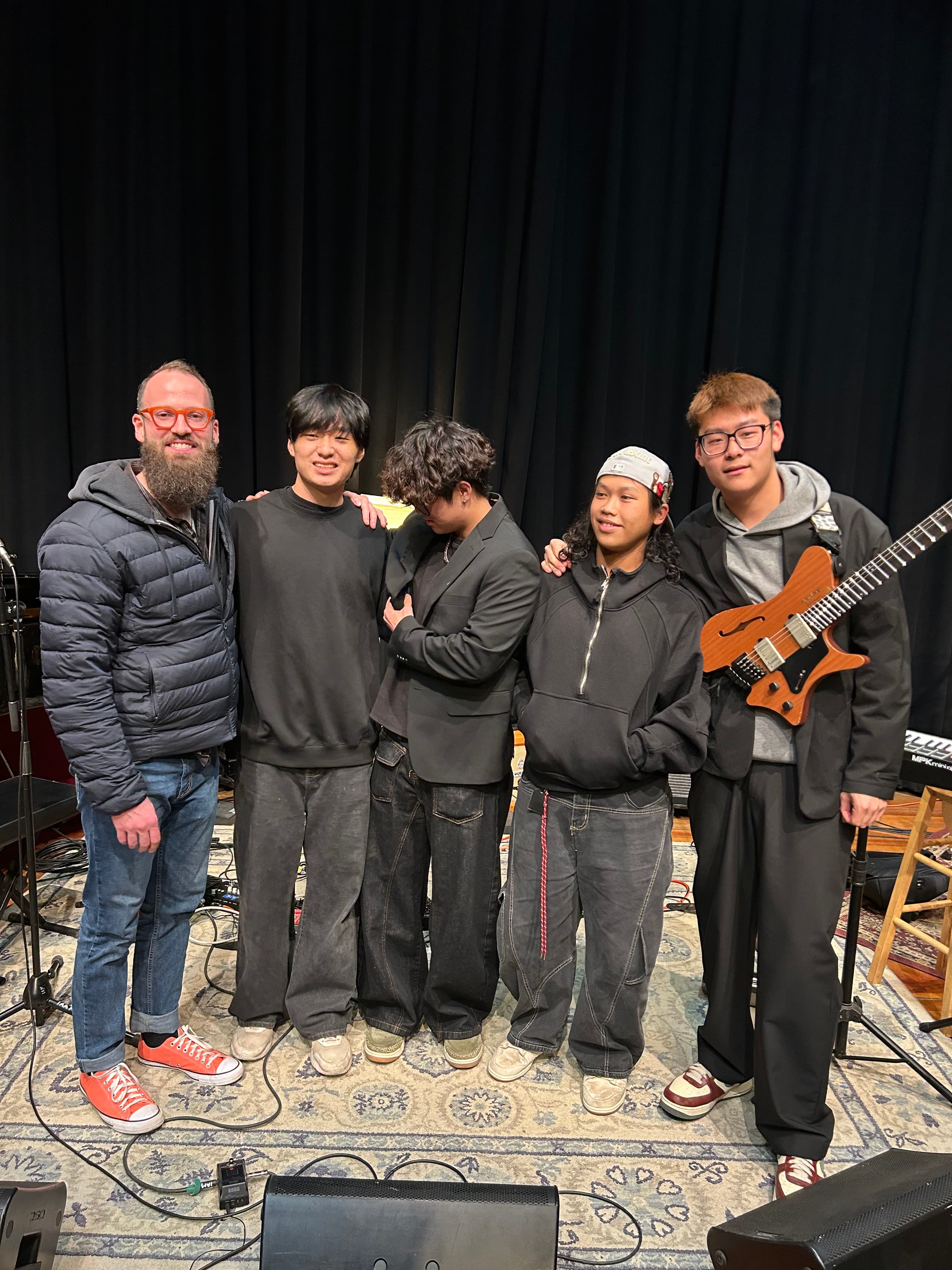 Five people pose together on a stage with a headless guitar and musical equipment.